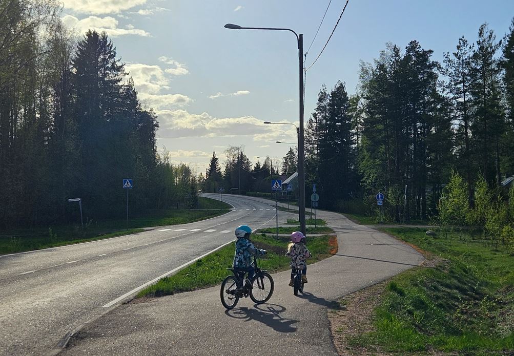 Kaksi pyöräilevää pikkulasta ylittämässä suojatietä ja jatkamassa matkaa jk pp-tiellä. 